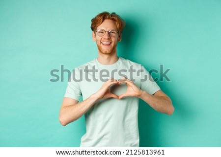 Valentines day and relationship concept. Happy boyfriend with red hair and beard, wearing glasses and t-shirt, showing heart sign and saying I love you, standing over turquoise background