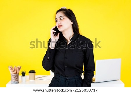Studio shot of Asian confused amazed doubt thoughtful female businesswoman employee in black outfit sitting on working desk checking news on e-mail from smartphone at office on yellow background.