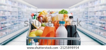 Shopping cart filled with food and drinks and supermarket shelves in the background, grocery shopping concept Royalty-Free Stock Photo #2125898954