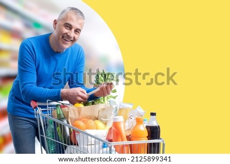 Smiling man leaning on a full shopping cart and checking the grocery receipt, offers and sale concept, blank copy space