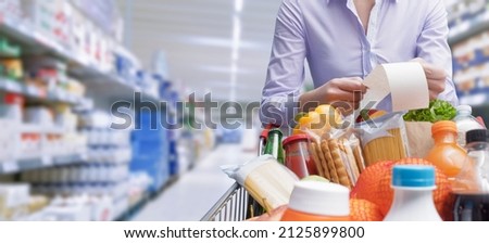 Woman pushing a cart and checking a grocery receipt, grocery shopping and expenses concept Royalty-Free Stock Photo #2125899800