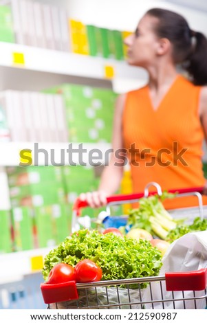 Young woman searching for products on the shelf with full trolley.