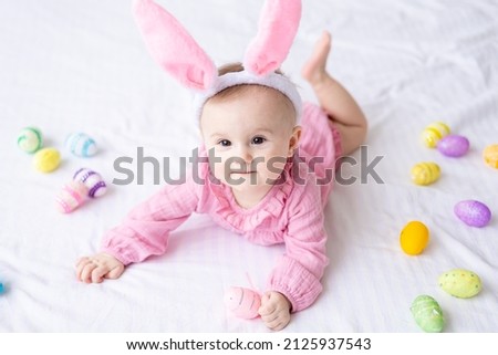 happy Caucasian baby girl with a rim of bunny ears on her head lies on the bed at home in the bedroom with colorful Easter eggs, easter baby