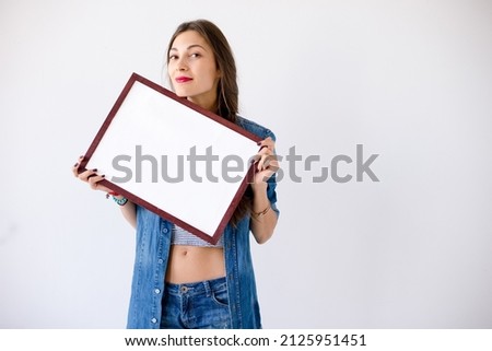 Playful girl dressed in denim hold a white(blink, empty) frame canvas in a white studio with copy space for product, graphic, video or text. Presentation, advertisement concept.