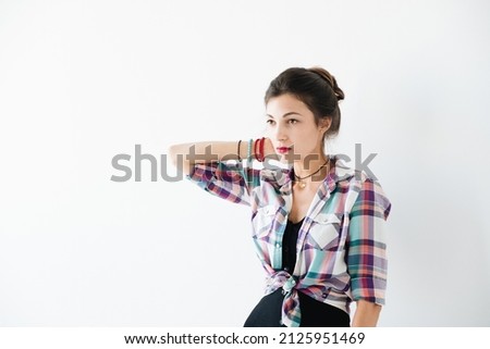 Worried tanned young woman wearing plaid shirt looks aside, with copy space above her head. Thoughtful girl holds one arm on her neck on white background. Worried about future, job problems.