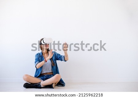 Smiling woman enjoy virtual reality with VR glasses gesticulating hands. Girl in denim shorts, shirt and boots, with crossed legs, use VR device while fly in cyberspace. Technology addicted.