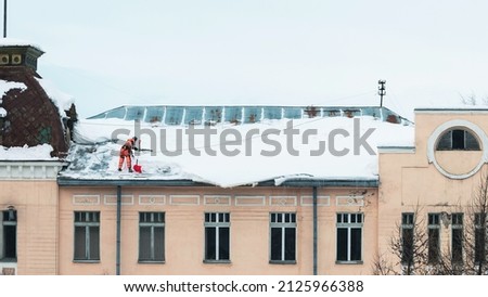 A working man in bright overalls with a safety belt with a shovel clears snow from the roof of an old building. Prevention of snow falling from the roof, industrial mountaineering