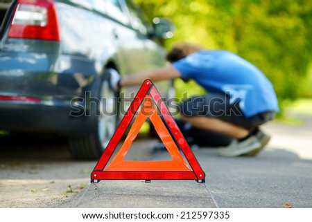 Red warning triangle sign on the road with a man checking his broken car in background