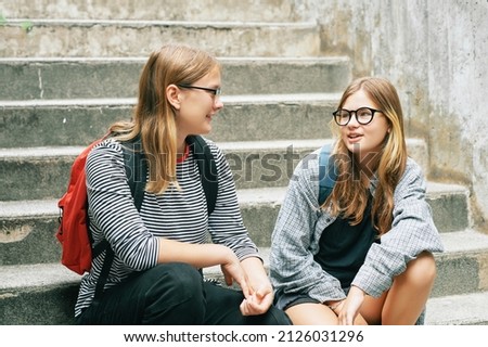 Outdoor portrait of two teenage girl sitting on stairs, wearing backbacks, talking to each other Royalty-Free Stock Photo #2126031296