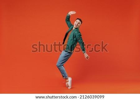 Full size body length smiling happy young brunet man 20s wears red t-shirt green jacket standing on toes dancing lean back have fun spreading hands isolated on plain orange background studio portrait.