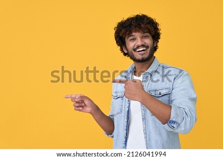 Happy excited young indian man looking at camera pointing aside with fingers hand gesture at copy space advertising new promotion, presenting sale offer standing isolated on yellow background.