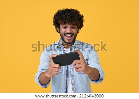 Excited happy young indian man gamer player using smartphone playing game in mobile videogame app or watching digital video stream holding cell phone isolated on yellow background.