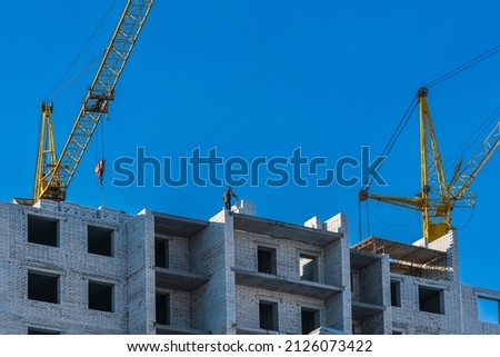 Two yellow construction cranes and a male builder on top of a house under construction against a blue sky. Bottom side view from below 