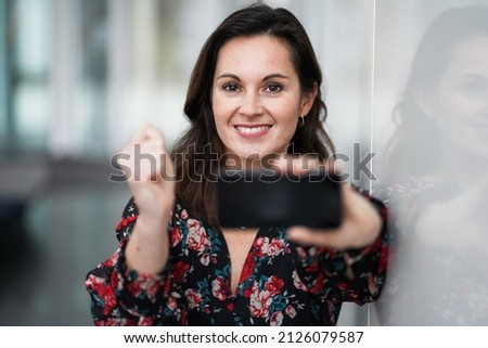 happy smiling chic business lady in red floral dress in a optimistic and cheerful mood holding and showing mobile phone into the camera pointing to the front
