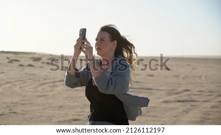 Young woman in desert taking photos or videos on mobile phone. Girl using smartphone. Sand storm, strong wind. Female photographer tourist traveler in Namibia, Africa.