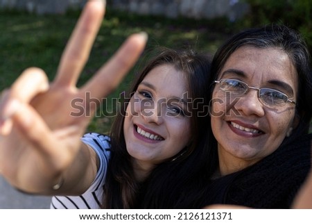 Latin American mother (50) and daughter (24) smile as they take a selfie and the daughter makes a peace sign with her hand. Mother's day concept and technology.