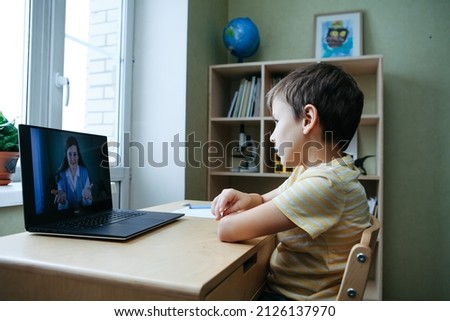7 years old boy sitting by desk with laptop doing writing task during online lesson. Side view. Smiling woman tutor on computer screen