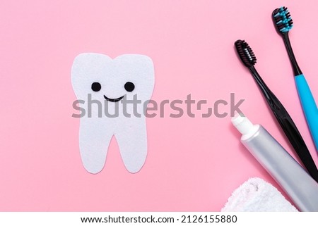 A silhouette of a tooth carved out of felt with a smiling cartoon face, next to an electric toothbrush, mouth freshener and toothpaste. Flat lay. Pink background. Oral hygiene concept.