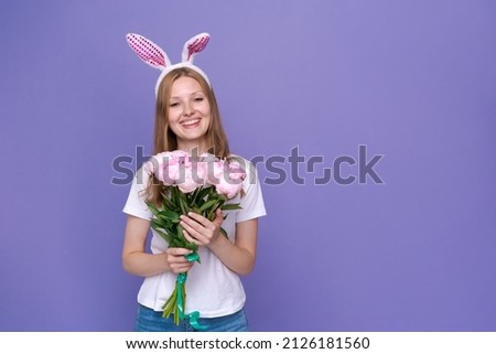 Easter. Happy attractive woman in pink easter bunny ears and white t-shirt, girl with a bouquet of beautiful peonies, on an isolated purple background. Caucasian girl rejoices in the spring holiday