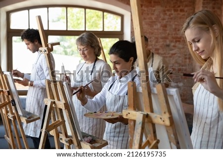 Happy focused diverse multiracial people in aprons using artist tools, enjoying painting group class drawing with paintbrushes on canvas, working together in modern studio, creative hobby activity. Royalty-Free Stock Photo #2126195735