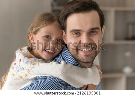 Happy millennial dad piggybacking cute pretty little preschool daughter kid, playing, carrying cheerful girl, looking at camera with toothy smile. Fatherhood concept. Headshot portrait