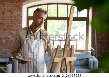 Relaxed happy middle aged 40s man in apron holding palette with mixed oil paints in hands, involved in painting picture on easel, enjoying creative hobby pastime alone in modern studio or at home.