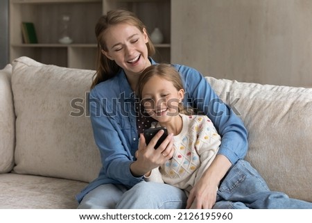 Cheerful young mother showing online learning app on smartphone screen to happy gen Z girl kid. Mom and cute little daughter using mobile phone together, shopping on Internet, making video call
