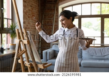 Happy inspired young Indian woman in apron and tied hair holding palette and paintbrush, creating new artwork on easel in modern gallery or studio. Joyful millennial female artist drawing picture.