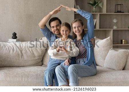 Happy family couple and cute child home portrait. Cheerful parents showing insurance, safety symbol, making house roof shape overhead, daughter kid showing hand heart love gesture, smiling at camera
