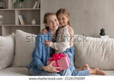 Happy touched young mom and cheerful cute daughter kid celebrating mothers day, birthday, 8 march, holding pink gift wrap, hugging on couch with love, tenderness looking at camera. Family portrait