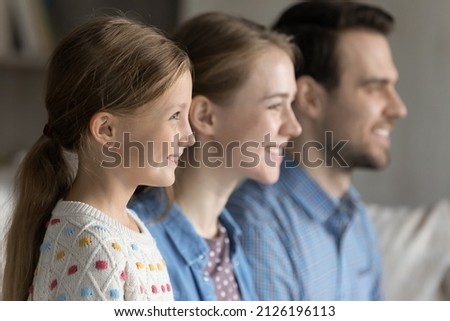 Happy pretty 7s girl kid home portrait, side view with mom and dad in blurred background. Family couple of parents and daughter girl looking away, smiling, sitting in line together