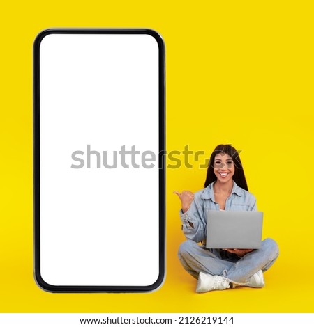 Great Offer. Excited lady sitting on floor using pc pointing finger at big giant cell phone with empty white screen, presenting new website, free copy space for mock up design, vertical banner