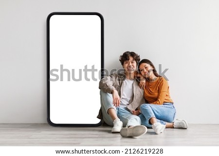Loving asian couple sitting near big cellphone with empty white screen, embracing, recommending newest mobile application or cool website over grey wall, mockup Royalty-Free Stock Photo #2126219228