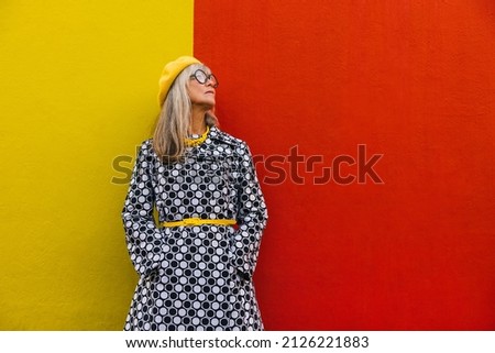 Elderly woman looking away thoughtfully while standing against a red background. Pensive senior woman reflecting on memories of the past. Stylish mature woman wearing colourful clothing. Royalty-Free Stock Photo #2126221883