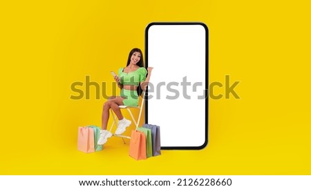 Mobile Shopping App. Excited Lady Sitting On Chair With Shopper Bags On Floor Pointing At Blank Empty Cell Screen Using Phone, Yellow Background. Online Order, Virtual Store Concept. Mock Up Banner