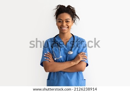 Portrait of positive female African American doctor or nurse posing with crossed arms and smiling looking at camera isolated over white studio background wall, lady wearing blue coat and stethoscope