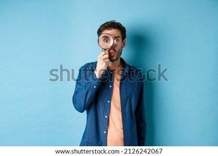 Image of surprised man found something interesting, say wow and look through magnifying glass, standing on blue background