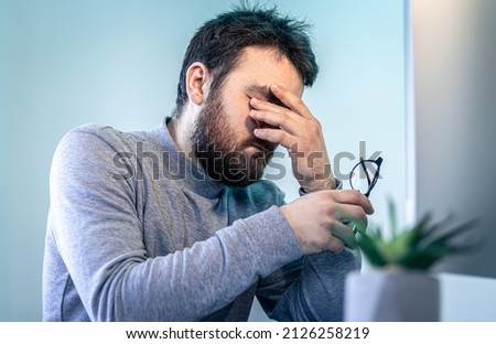 A tired man rubs his eyes in front of a computer screen.