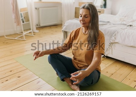 Indoor picture of sporty mature female doing mudra sign with hands, breathing deeply, mediating keeping eyes closed sitting on floor on green mat against bedroom background with bed, window and mirror