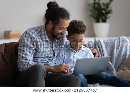 Caring happy young African American father helping small cute kid son using computer software application, playing entertaining video games, shopping in internet store, tech addiction concept.