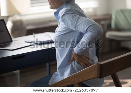 Cropped unhealthy middle aged senior woman employee massaging stiff back muscles, suffering from sudden pain, sitting at table doing computer work, feeling strong lumbar ache at modern home office. Royalty-Free Stock Photo #2126337899