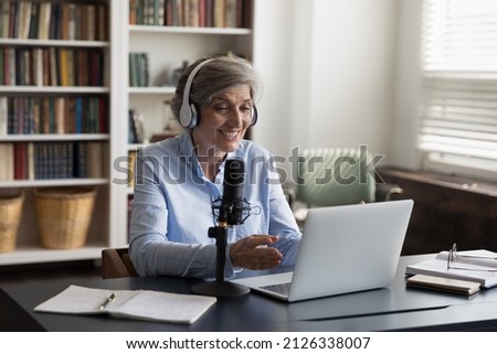 Smiling motivated beautiful old mature woman in headphones speaking in professional stand mic, voice acting, recording educational video or streaming online, elderly people and modern tech usage.
