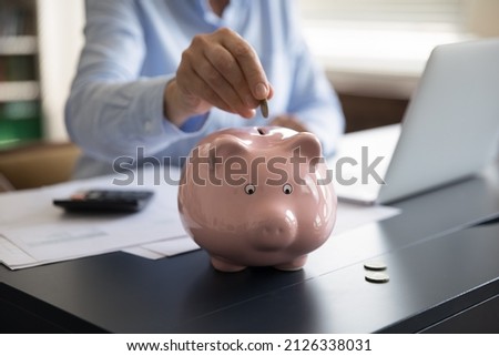 Close up cropped middle aged mature woman putting coin in small piggybank, saving money for healthcare medical insurance, planning investment, managing household budget, sitting at table alone at home Royalty-Free Stock Photo #2126338031