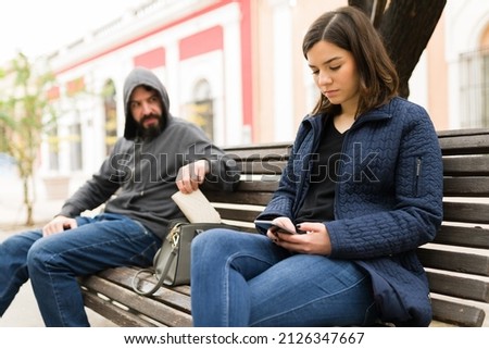 Attractive young woman texting while sitting on a bench on the street next to a thief pickpocketing her purse Royalty-Free Stock Photo #2126347667