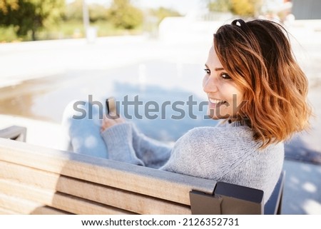 Cute pretty young woman with a charismatic grin relaxing on a wooden banch in a city park turning to look to the side with a beaming smile Royalty-Free Stock Photo #2126352731