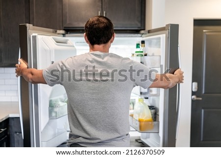 Back of hungry man opening fridge refrigerator doors domestic appliance searching for food inside with condiments and juice in modern kitchen Royalty-Free Stock Photo #2126370509