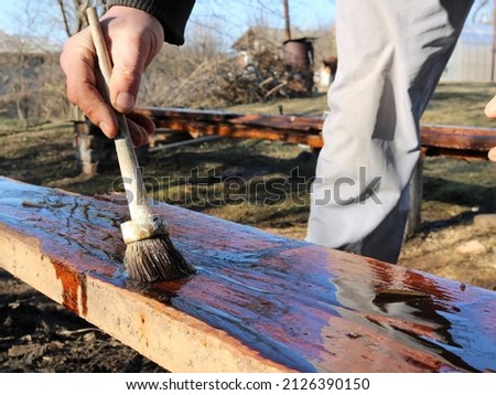 application of protective oil on the surface of wood in rustic construction, a man impregnates a wide board with technical liquid on a sunny street, coating with oil to protect wood material Royalty-Free Stock Photo #2126390150