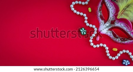 Masquerade mask, yellow feathers, gummies and white pearl beads lie on the right on a red background with copy space on the left, flat lay close-up.Mardi gras holiday concept.