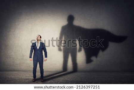 Determined businessman stands confident in a hero stance and casting a brave superhero shadow on the wall behind. Business leadership and motivation concept. Ambition and strength symbols Royalty-Free Stock Photo #2126397251