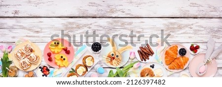 Easter breakfast or brunch bottom border. Top view on a white wood banner background. Bunny pancake, egg nests, chick fruit and an assortment of spring food items. Copy space. Royalty-Free Stock Photo #2126397482